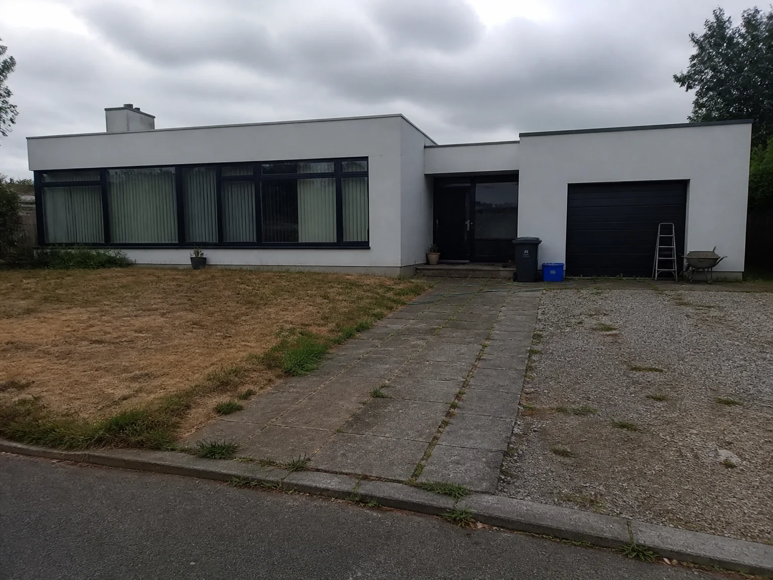Casa moderna de telhado plano · céu cinzento, relvado seco, cascalho com ervas daninhas e pavimento, contentores, escada e carrinho de mão — tudo corrigido, a casa inalterada — before